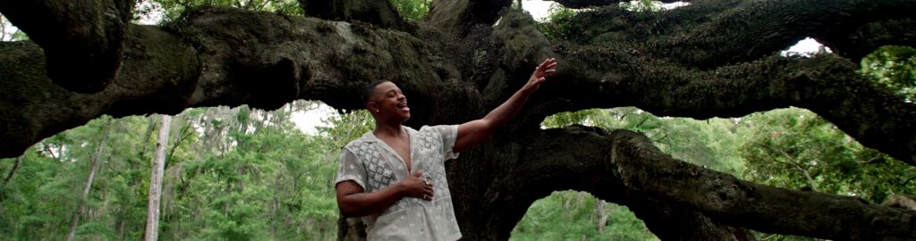 A young Black man in a white lace shirt raises his hand towards the sky as he sings. He stands in front of a large oak tree with many thick branches.