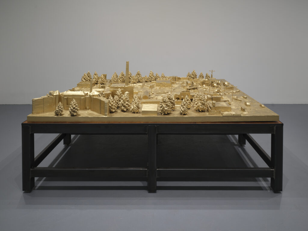 Bronze cast models of a city landscape with trees, billboard, smokestacks, armored police cars, and telephone poles atop metal tables.