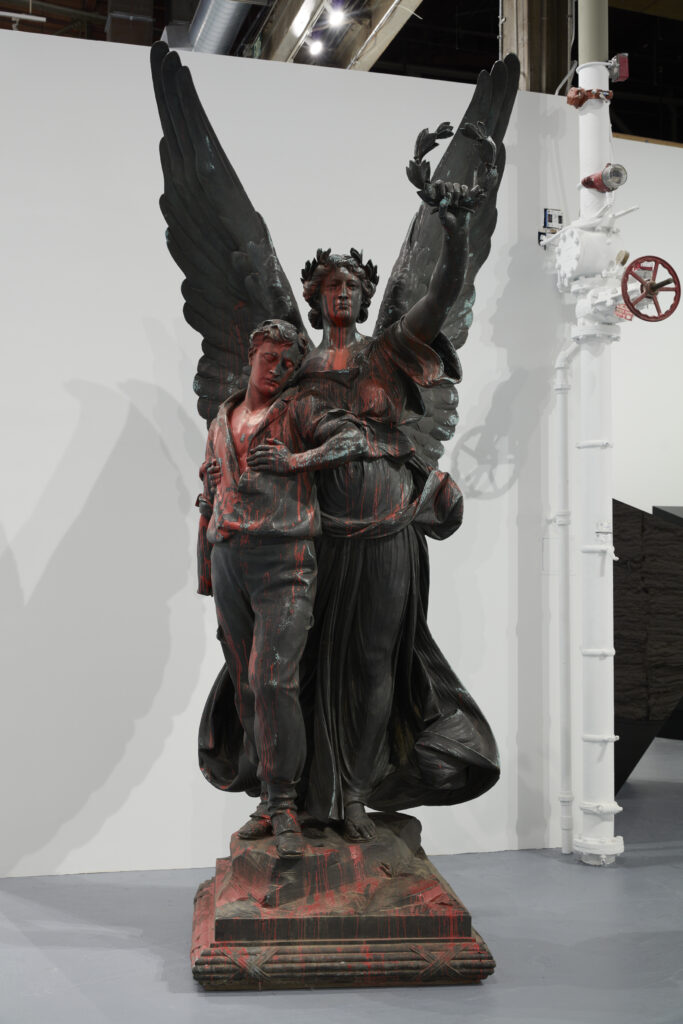 Bronze statue of winged female allegorical figure holding a dying young soldier in one arm and holding up a laurel wreath with the other. Red paint has been splashed on the monument