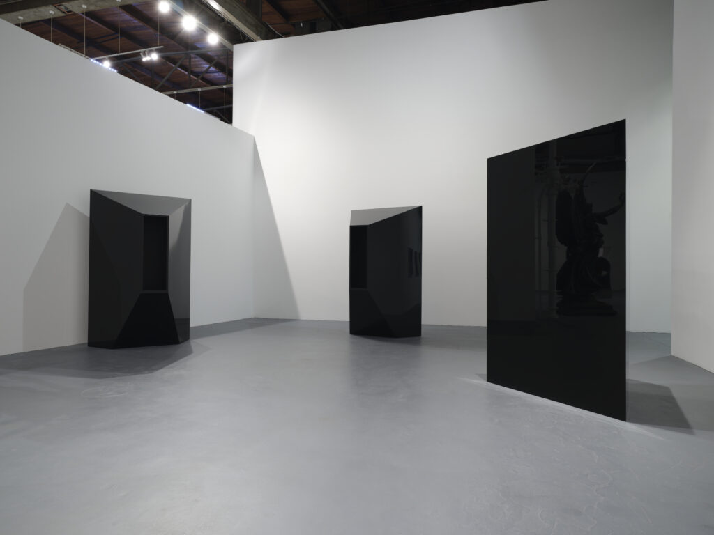 Three reflective black trapezoidal sculptures stand in a triangular arrangement