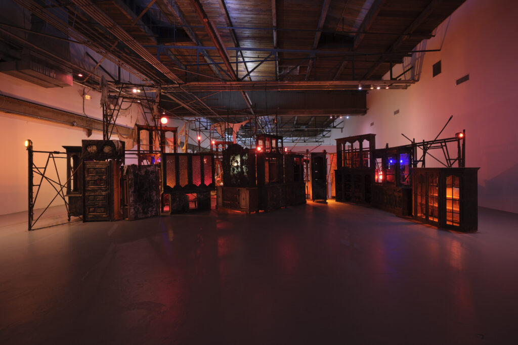 A mass of wooden china cabinets and scaffolding covered in singed fabric with glowing orange, red, and blue lights.
