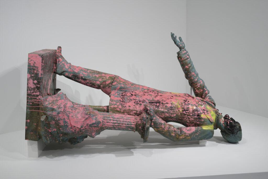 A bronze statue that has been splattered with yellow and pink paint. It lays on its back with its arm outstretched to the sky. The face and head have been flattened.