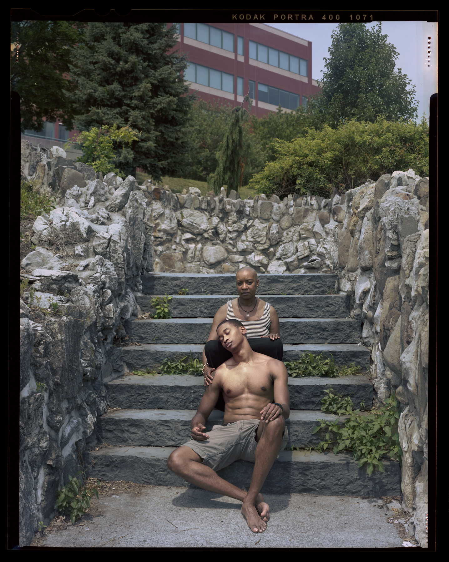 A Black mother sits on a set of stone steps looking at the camera. Her shirtless teen son sits below her, leaning against her lap with his eyes closed. An office building surrounded by trees is visible in the background